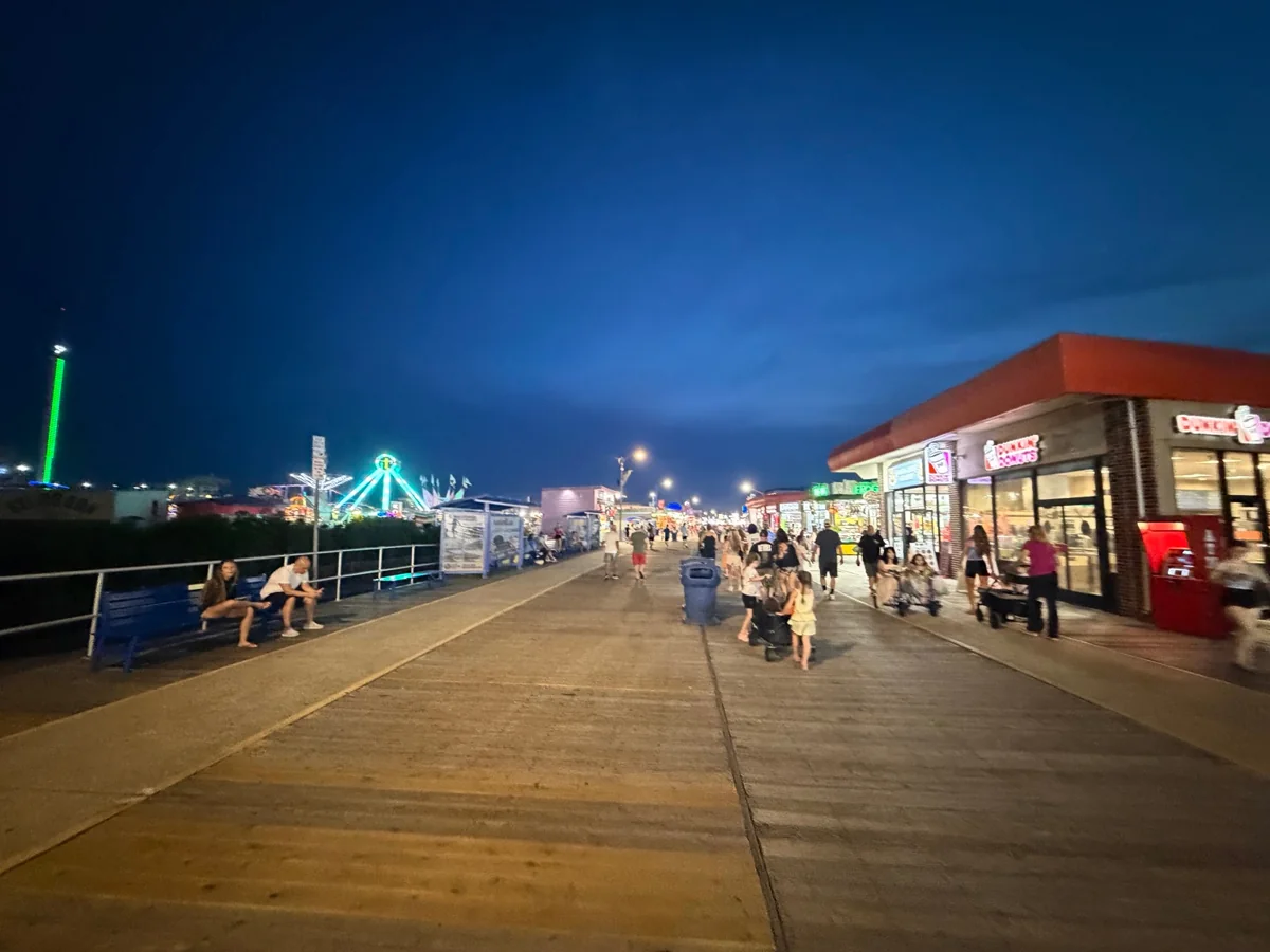 A Wildwood boardwalk evening with glowing rides, families strolling and a deep-blue sky