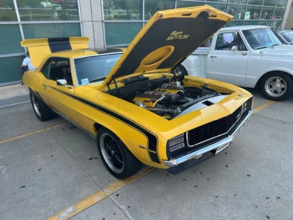 A yellow classic sports car with black stripes and its hood open at a car show