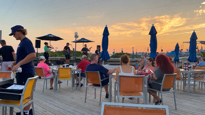Musicians playing on an outdoor deck at Seaport Pier at sunset, with blue umbrellas and dining tables